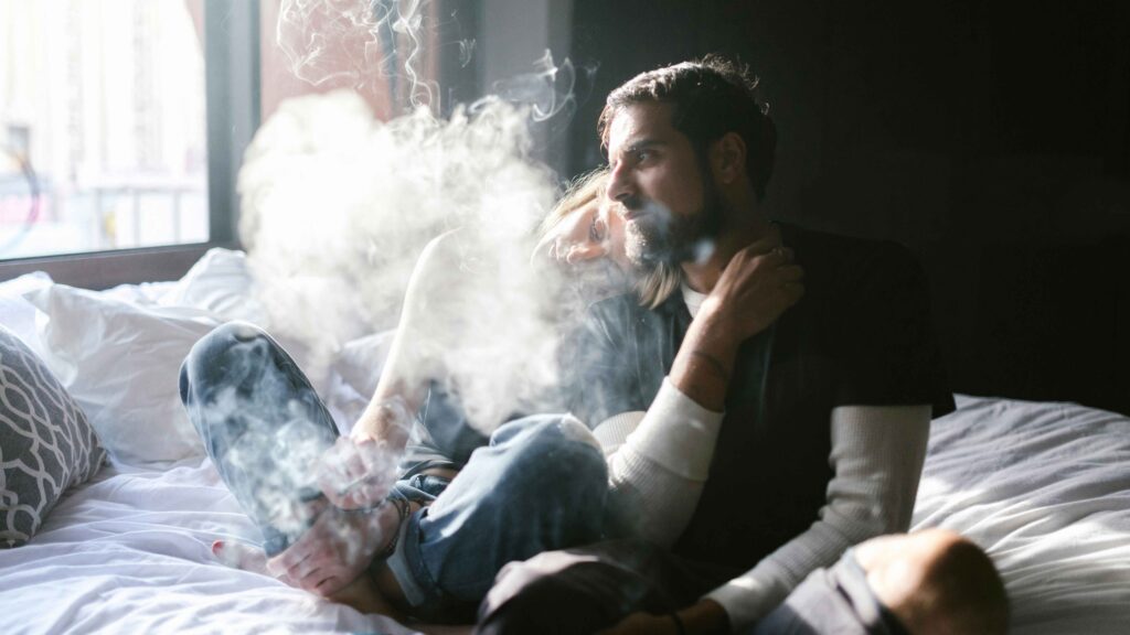 Cigarettázó férfi ül az ágy szélén, párja a vállára dől; füsttel teli, intim hangulatú beltéri jelenet.
