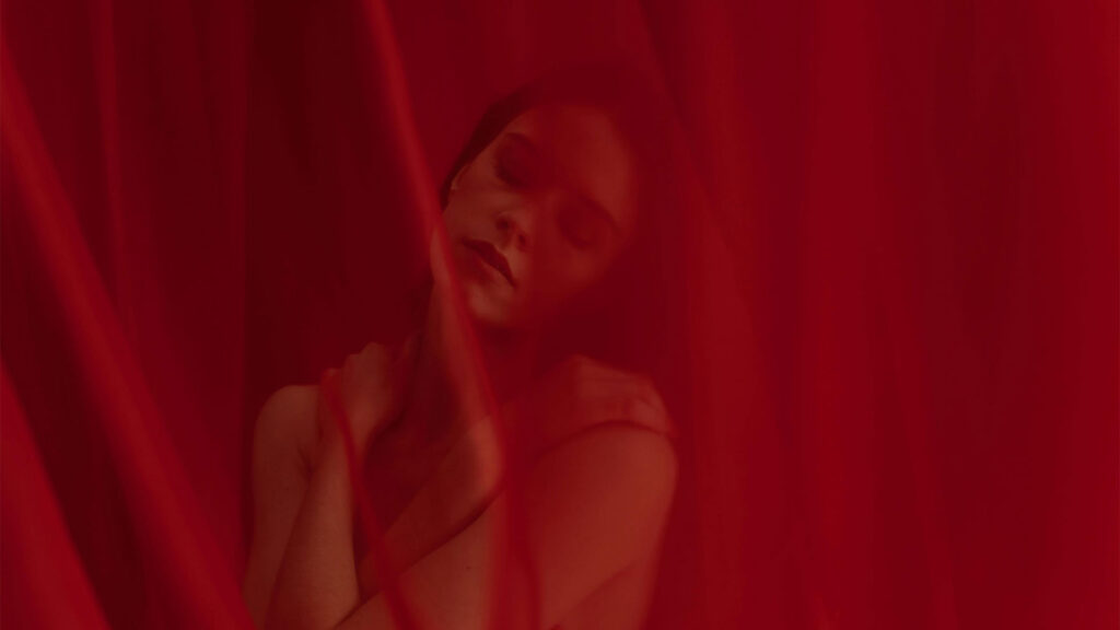 Fiatal nő csukott szemmel áll áttetsző vörös fátyol mögött és a vállát érinti miközben nyugodt intim hangulatú portré készül róla az endometriózis illusztrálására.