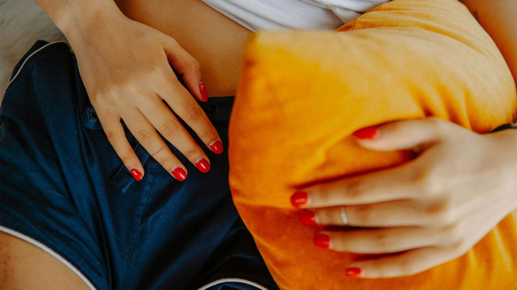 Nő párnába kapaszkodva fekszik az ágyon és hasi diszkomfortot valamint menstruációs fájdalmat él át.