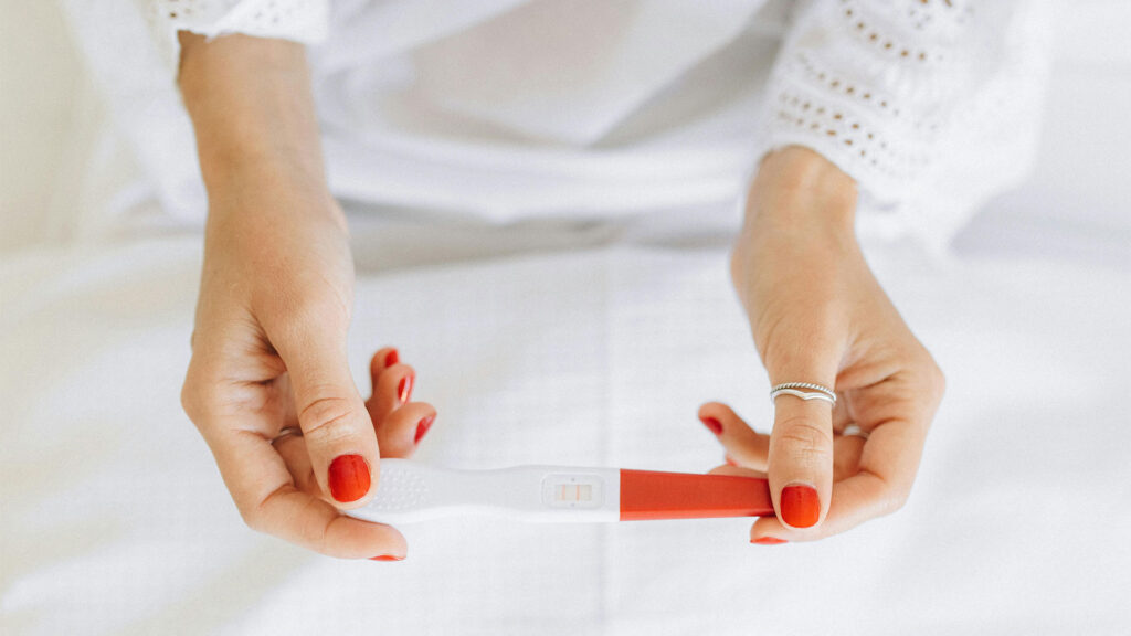 Női kezek pirosra lakkozott körmökkel egy pozitív terhességi tesztet tartanak fehér ágynemű fölött, a teszten két csík látható.