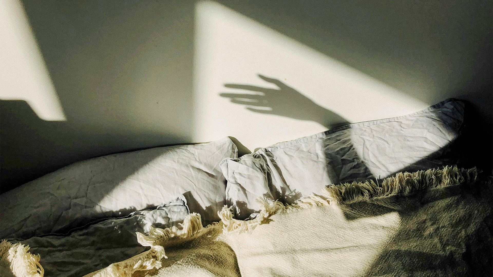 Napfényes hálószoba ágyban fekvő párnákkal és falra vetülő kézárnyékkal, nyugodt esti hangulatban.
