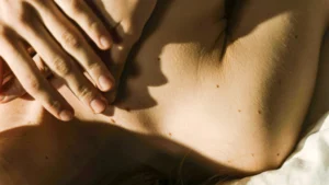 Közeli felvétel egy nő dekoltázsáról és nyakáról, finoman a bőréhez érő kézzel, meleg fényben.